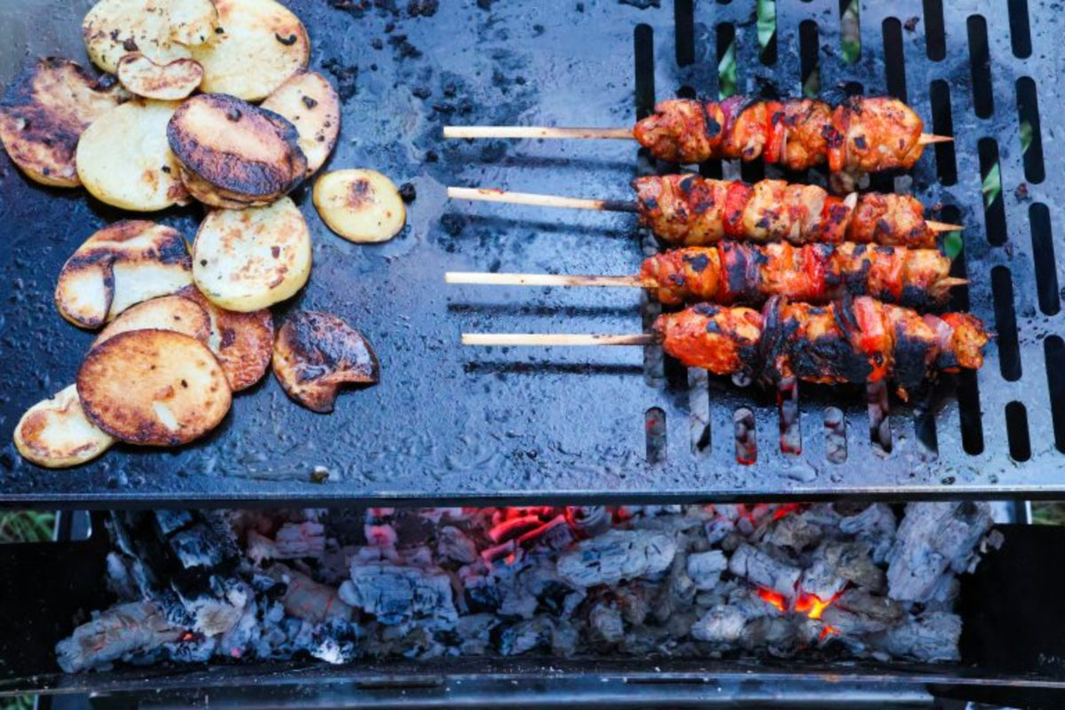 Portable camping fire pit cooking skewers and potatoes on BBQ plate over coals